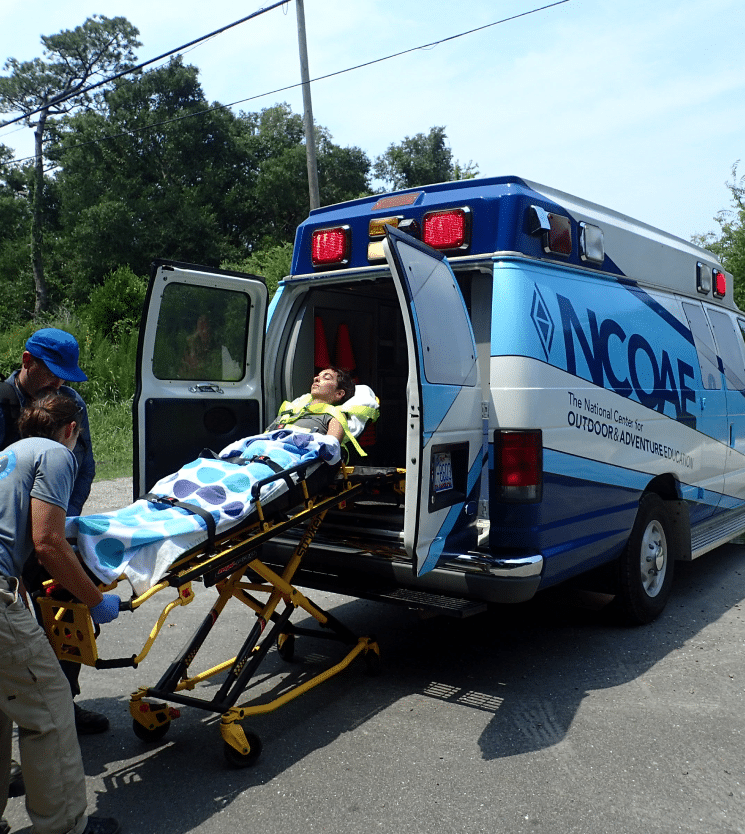 A person on a stretcher being pushed by an EMT into an NCOAE vehicle.
