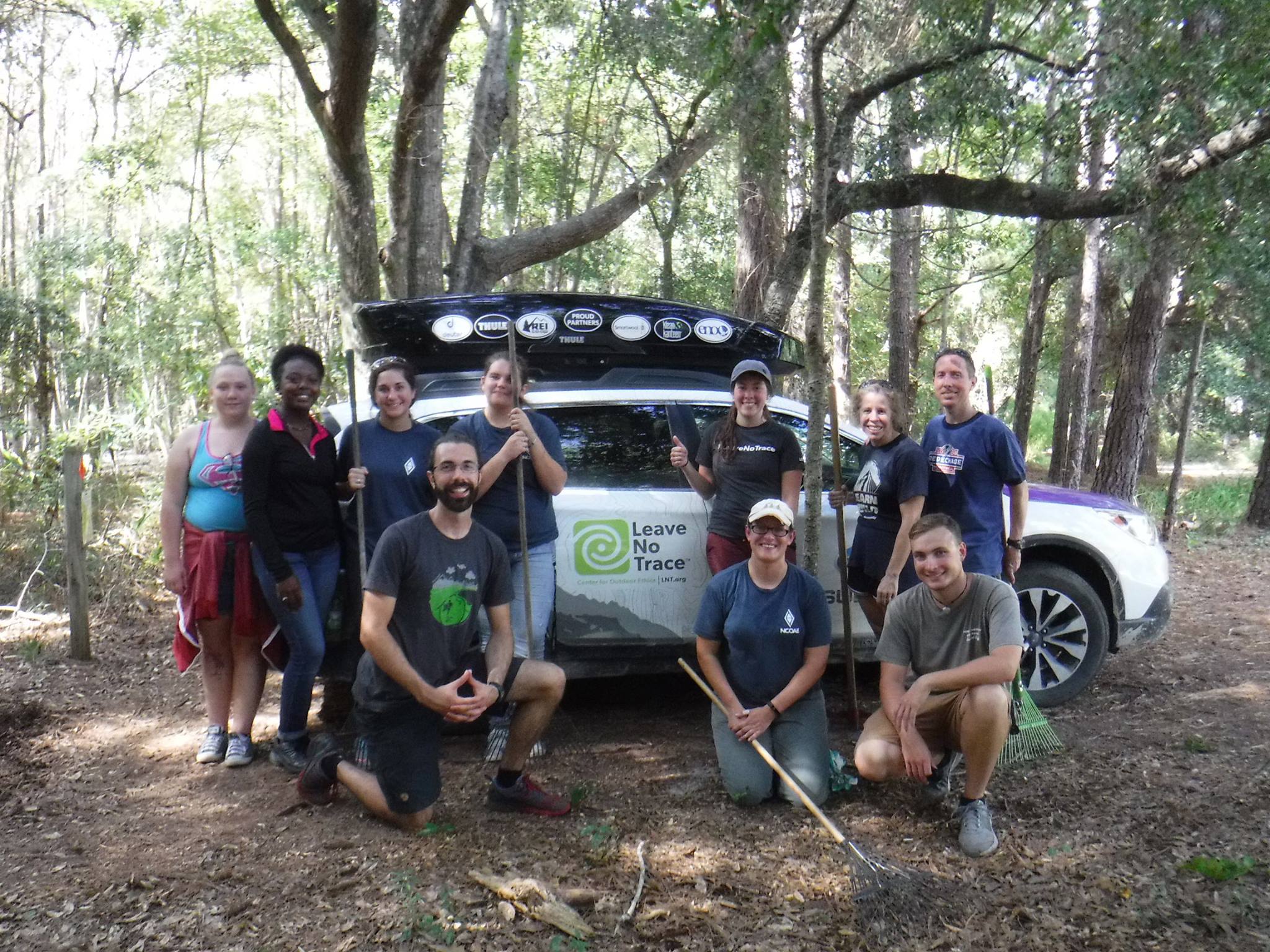 A group of people posing in front of a car in the woods.