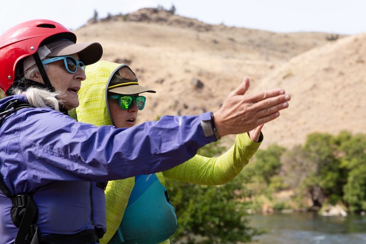 A woman wearing a helmet and a pair of goggles is pointing to the river.