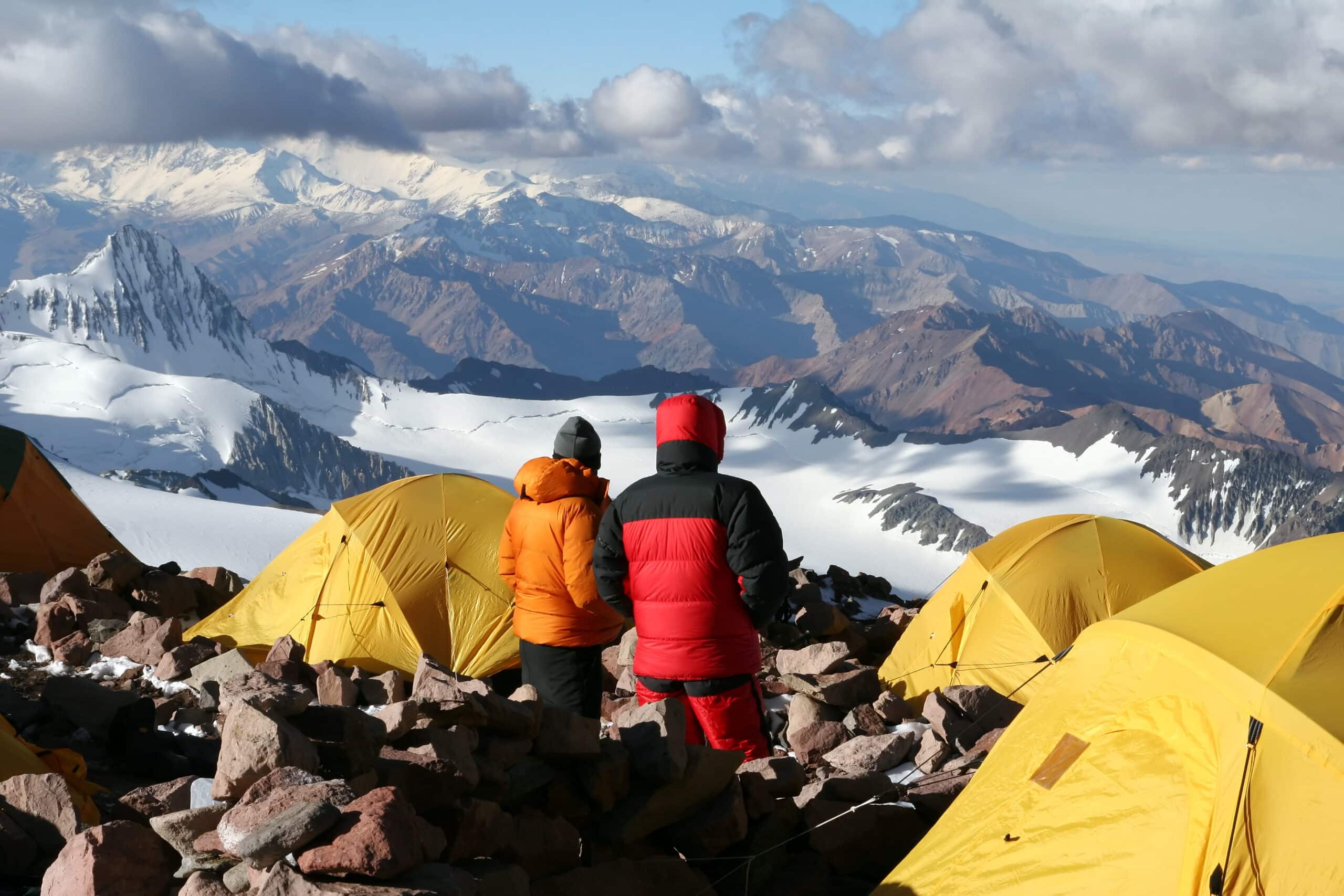 Two people standing on top of a mountain with tents in the background.