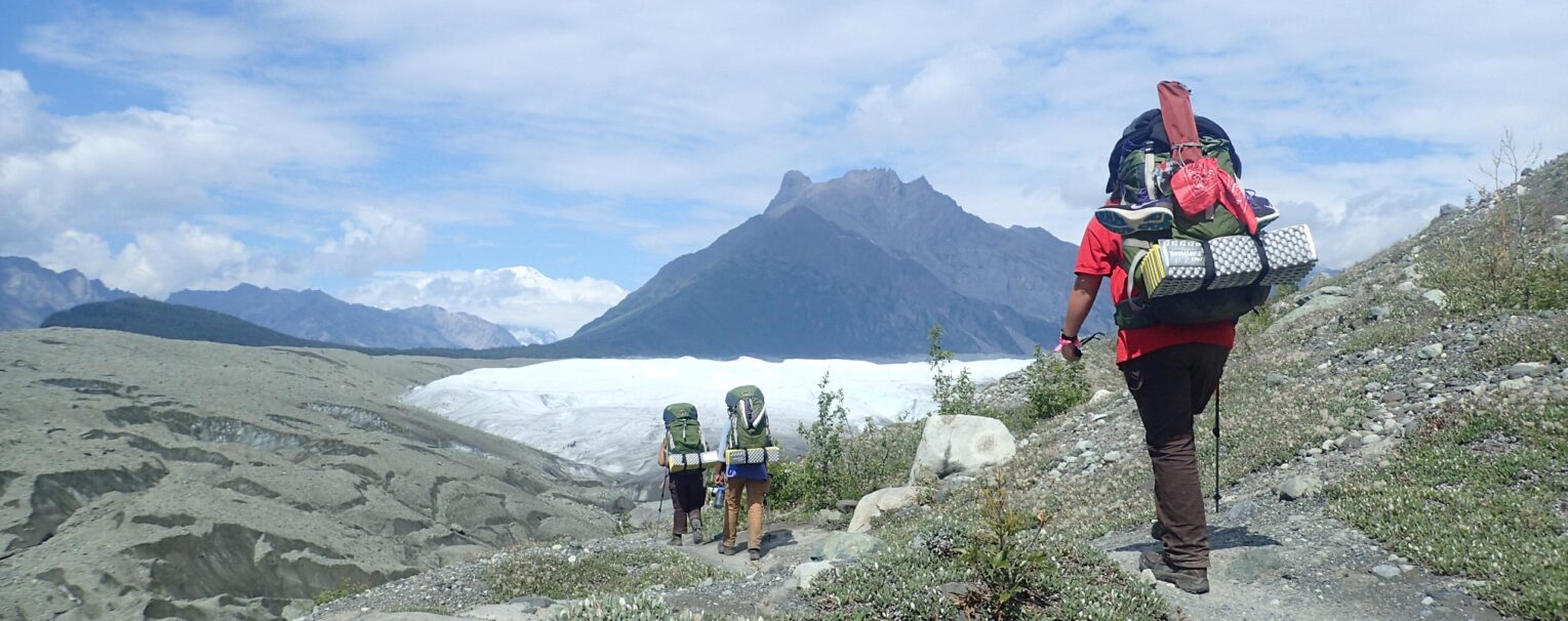 Three people backpacking in Alaska with a view of a mountain ahead of them.