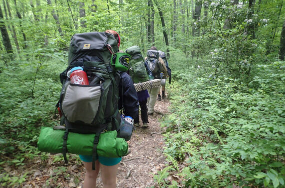 A group of people wearing and carrying outdoor gear while hiking in a forest.