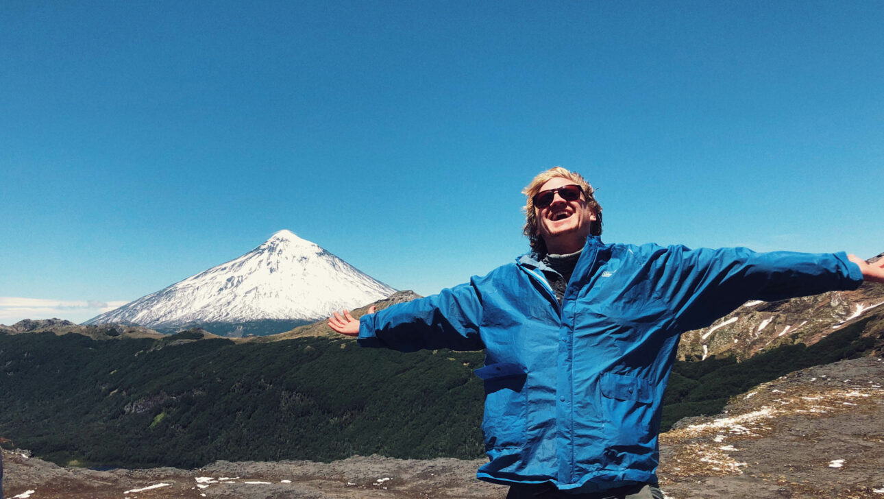 A person posing with their arms outstretched with a view of a snowcapped mountain in the background.