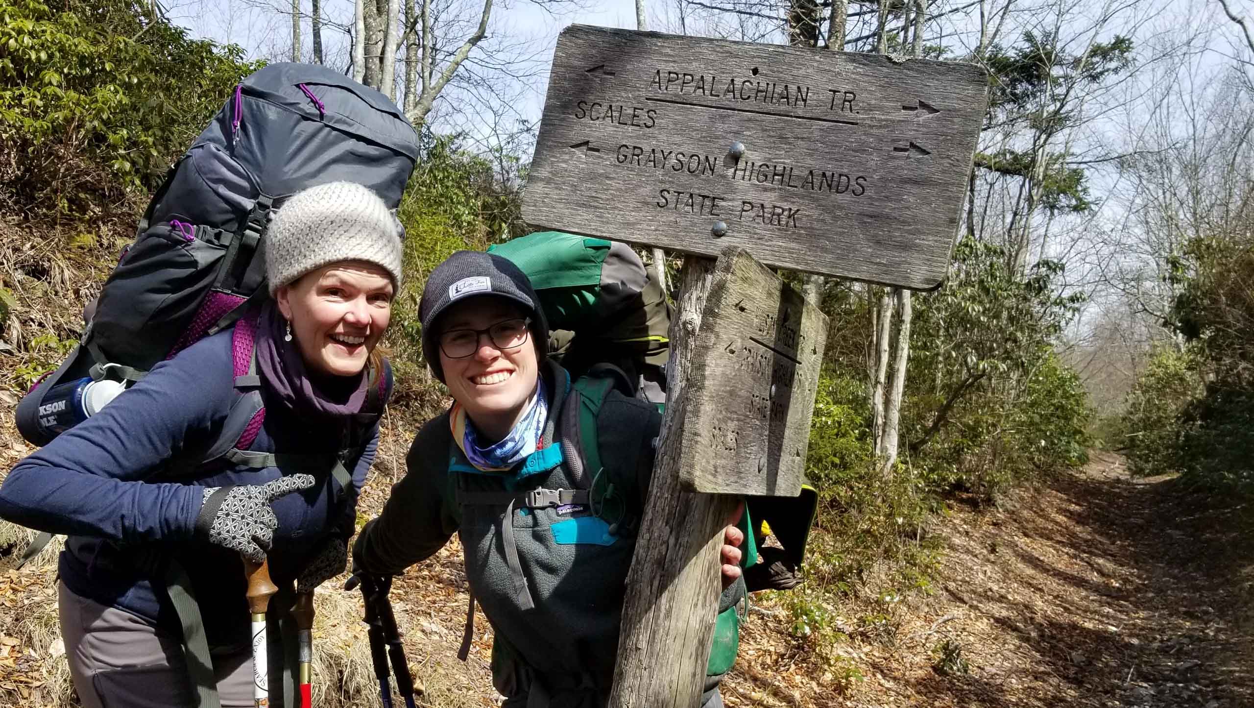 Two women with hiking gear, smiling next to a sign on a trail.