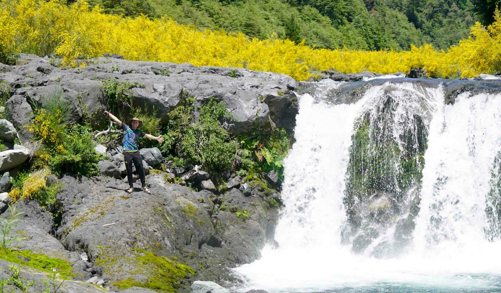 A person posing with their arms outstretched next to a waterfall.