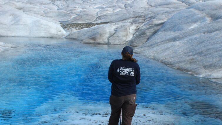 A person wearing an NCOAE t-shirt, standing in front of glaciers.