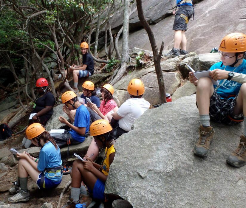 A group of students wearing rock climbing gear, taking notes in their journals.