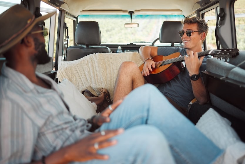 Smiling guys sitting in the back of a van playing guitar on a road trip