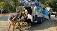 Two people assist a woman onto a stretcher and into an ambulance parked on a gravel surface with trees in the background.