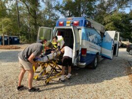 Two people assist a woman onto a stretcher and into an ambulance parked on a gravel surface with trees in the background.